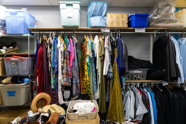 Brightly coloured dresses and shirts items on racks labeled “Women’s light tops” and “Mens heavy tops” surrounded by plastic storage bins overflowing with hats, shoes and other clothing items in the back room of a Winnipeg thrift store