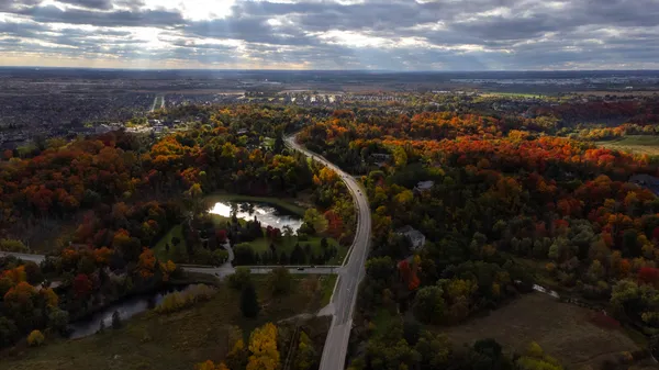 Overhead view of a roadway weaving through a forested area in the fall, with sun peeking through the clouds
