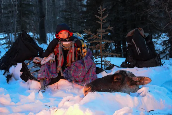 A young child waiting by the fire as his dad finishes packing up a caribou